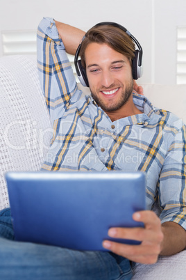 Casual smiling man lying on couch listening to music on tablet p