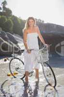 Beautiful blonde in white sundress standing with bike on the bea