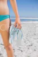 Mid section of fit woman in bikini on beach holding flip flops