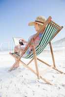 Woman in sunhat sitting on beach in deck chair using tablet pc