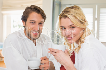 Cute couple in bathrobes having coffee together smiling at camer