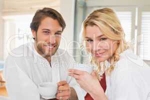 Cute couple in bathrobes having coffee together smiling at camer