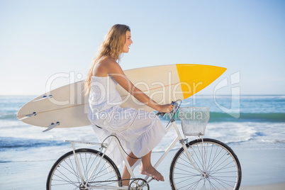 Beautiful surfer in sundress on bike holding surfboard at the be