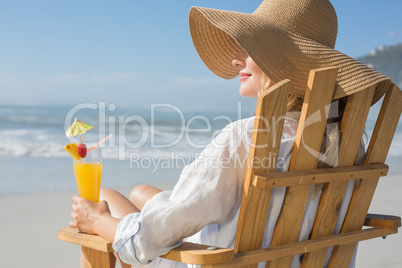 Smiling blonde relaxing in deck chair by the sea holding cocktai