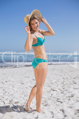 Slender woman in bikini on beach wearing sunhat smiling at camer