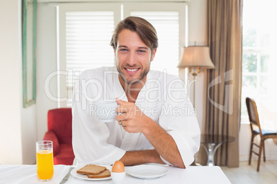 Handsome man having breakfast in his bathrobe smiling at camera