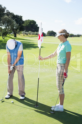 Lady golfer holding eighteenth hole flag for partner putting bal