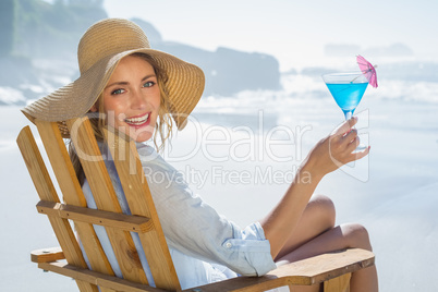 Smiling blonde relaxing in deck chair by the sea holding cocktai
