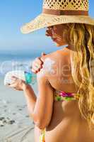 Gorgeous woman sitting on the beach in sunhat applying suncream