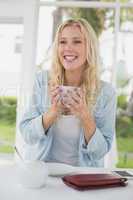 Pretty blonde sitting at table having coffee smiling at camera