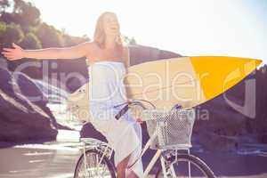 Beautiful surfer in sundress on bike holding surfboard at the be