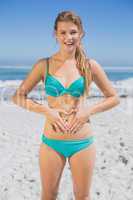 Fit smiling woman in bikini on the beach making heart shape on s