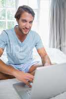 Handsome smiling man sitting on bed using laptop