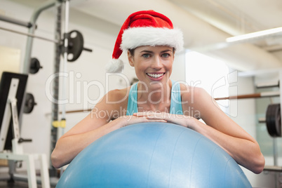 Fit smiling brunette in santa hat leaning on exercise ball