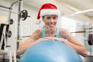 Fit smiling brunette in santa hat leaning on exercise ball