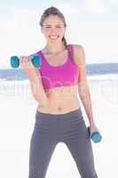 Fit woman working out with dumbbells on the beach smiling at cam