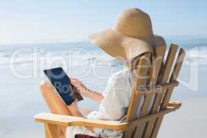 Smiling blonde sitting on wooden deck chair by the sea using tab