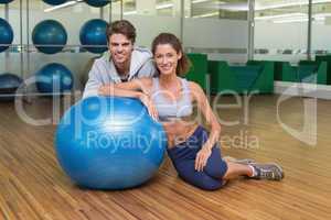 Fit woman leaning on exercise ball with trainer smiling at camer