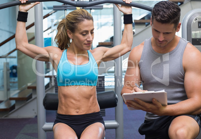 Female bodybuilder using weight machine for arms with trainer ta