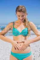 Fit smiling woman in bikini on the beach making heart shape on s