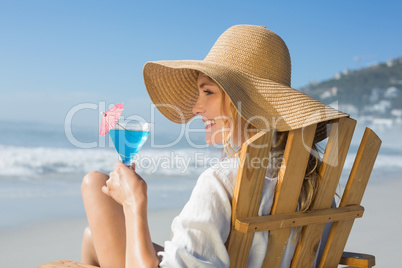 Smiling blonde relaxing in deck chair by the sea holding cocktai