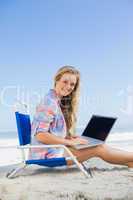 Pretty blonde sitting on beach using her laptop smiling at camer