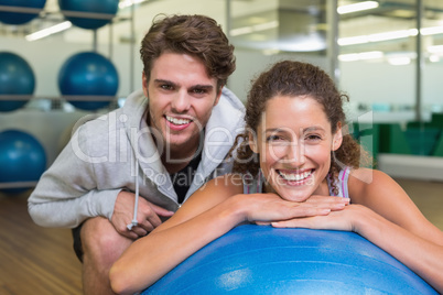 Fit woman leaning on exercise ball with trainer smiling at camer