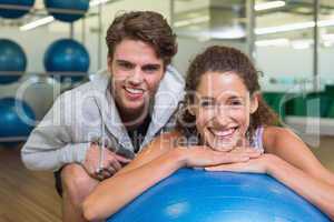 Fit woman leaning on exercise ball with trainer smiling at camer