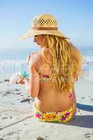 Gorgeous woman sitting on the beach in sunhat applying suncream