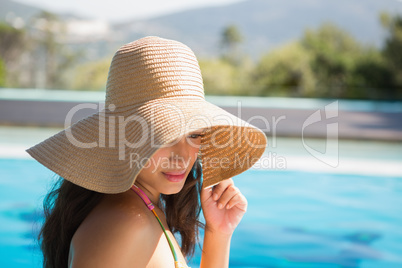 Beautiful brunette sitting by pool wearing straw sunhat