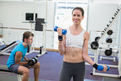 Fit brunette exercising with blue dumbbells smiling at camera