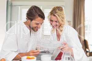 Cute couple in bathrobes having breakfast together reading the n