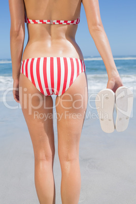 Mid section of fit woman in bikini on the beach holding flip flo