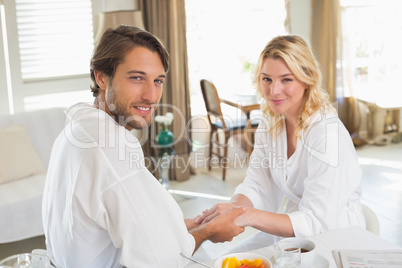 Cute couple in bathrobes having breakfast together holding hands