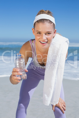 Sporty smiling blonde standing on the beach with towel and bottl