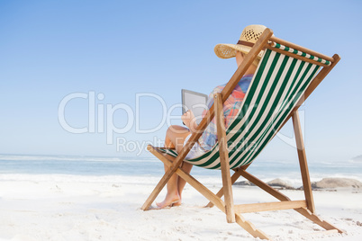 Woman in sunhat sitting on beach in deck chair using tablet pc