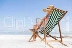 Woman in sunhat sitting on beach in deck chair using tablet pc