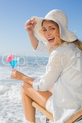 Smiling blonde relaxing in deck chair by the sea holding cocktai