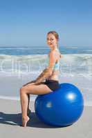 Fit woman sitting on exercise ball at the beach smiling at camer