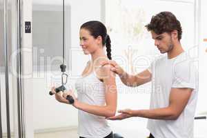 Fit smiling woman using weights machine for arms with her traine