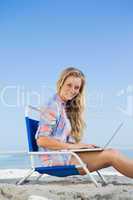 Pretty blonde sitting on beach using her laptop smiling at camer