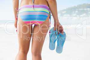 Lower half of fit woman holding flip flops on beach