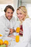 Cute couple in bathrobes having breakfast together smiling at ca