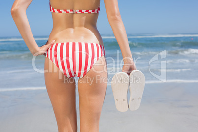 Mid section of fit woman in bikini on the beach holding flip flo