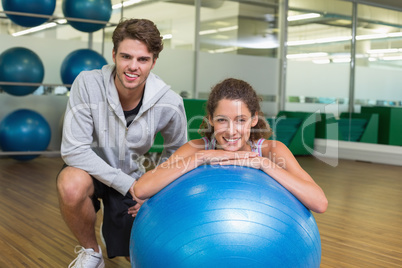 Fit woman leaning on exercise ball with trainer smiling at camer