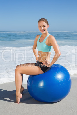 Fit woman sitting on exercise ball at the beach smiling at camer