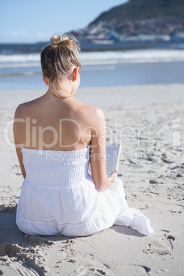 Content blonde in white dress sitting on the beach reading book