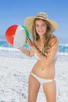 Fit blonde in white bikini and straw hat holding beach ball