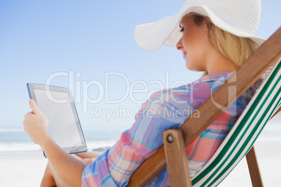 Woman in sunhat sitting on beach in deck chair using tablet pc