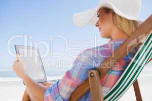 Woman in sunhat sitting on beach in deck chair using tablet pc